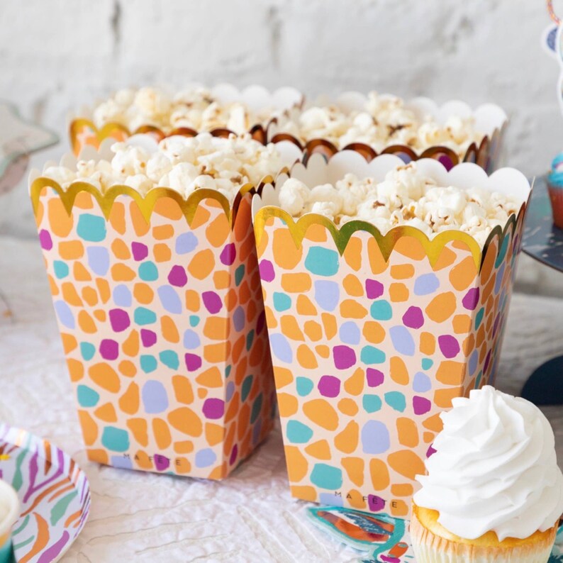 May include: Three popcorn containers with a colorful geometric pattern and gold trim. The containers are filled with popcorn and are sitting on a white surface.