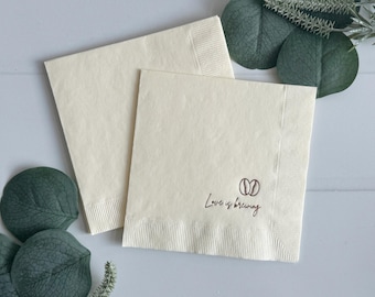 Peut inclure: Deux serviettes en papier crème. L'une est posée sur l'autre. Le coin inférieur droit de la serviette supérieure porte les mots "Love is brewing" et un motif de grains de café marron.
