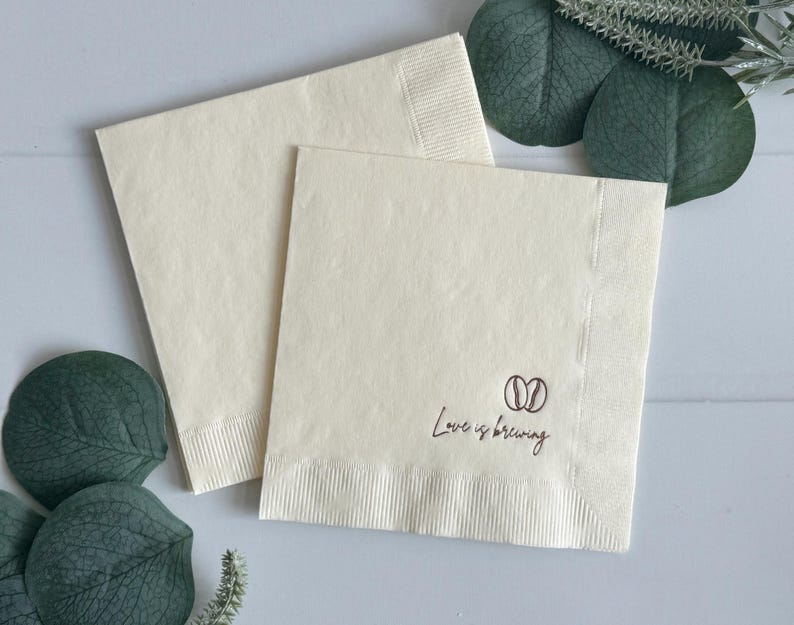 May include: Two cream-colored paper napkins. One napkin is on top of the other. The bottom right corner of the top napkin has the words "Love is brewing" and a coffee bean graphic printed in brown.