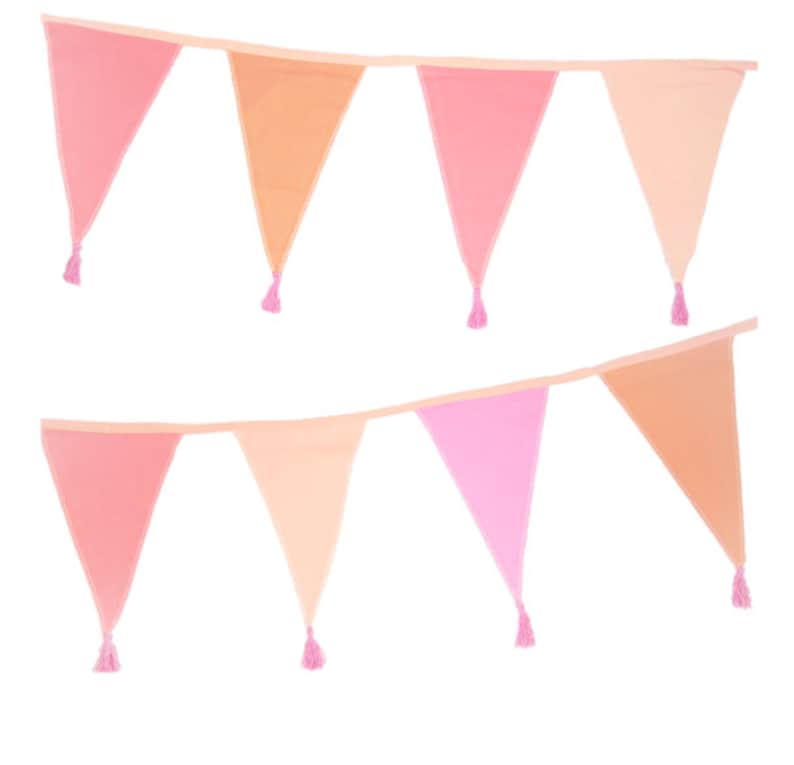 May include: A decorative bunting banner featuring two rows of triangular fabric flags in various shades of pink, peach, and coral. Each individual flag is finished with a small, matching pink tassel attached to the bottom point.