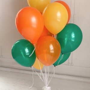 Op de afbeelding: Een boeket kleurrijke ballonnen in tinten van oranje, geel en groen. De ballonnen zijn samengebonden met wit lint, wat een feestelijke opstelling cre&euml;ert. De ballonnen zijn een mix van effen en doorschijnende kleuren.