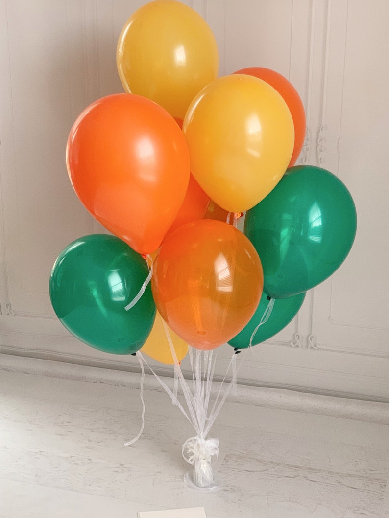 Op de afbeelding: Een boeket kleurrijke ballonnen in tinten van oranje, geel en groen. De ballonnen zijn samengebonden met wit lint, wat een feestelijke opstelling cre&euml;ert. De ballonnen zijn een mix van effen en doorschijnende kleuren.