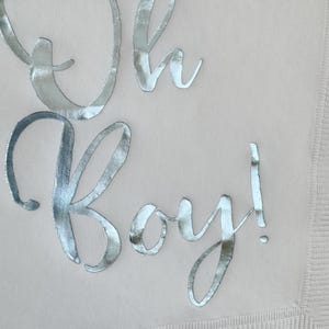 May include: A light beige paper napkin featuring the text Oh Boy! written in a reflective silver metallic script font. The napkin has a textured border along the edge, providing a subtle contrast to the smooth surface where the lettering is placed.
