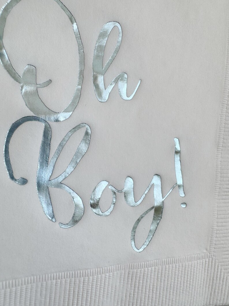 May include: A light beige paper napkin featuring the text Oh Boy! written in a reflective silver metallic script font. The napkin has a textured border along the edge, providing a subtle contrast to the smooth surface where the lettering is placed.