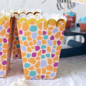 K&ouml;nnte beinhalten: Popcorn-Boxen mit einem farbenfrohen Terrazzo-Muster in Orange, Lila, T&uuml;rkis und Hellblau. Die Boxen haben einen goldfarbenen, gewellten Rand und sind mit Popcorn gef&uuml;llt. Der Text "MA FETE" ist auf die Vorderseite der Box gedruckt.