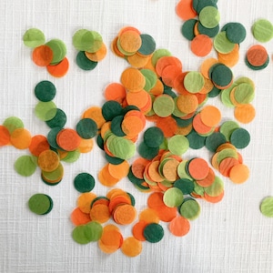 Op de afbeelding: Een verspreiding van cirkelvormige confetti in groen-, oranje- en donkergroentinten. De confetti ligt op een wit oppervlak. De confetti lijkt van dun papier te zijn gemaakt.