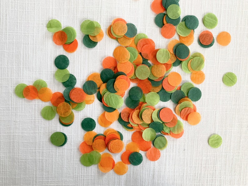 Op de afbeelding: Een verspreiding van cirkelvormige confetti in groen-, oranje- en donkergroentinten. De confetti ligt op een wit oppervlak. De confetti lijkt van dun papier te zijn gemaakt.