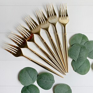 Op de afbeelding: Een bovenaanzicht van goudkleurige vorken in een waaier, vergezeld van groene eucalyptusbladeren. De vorken hebben een klassiek ontwerp met tanden en lange, slanke handvatten. De opstelling staat op een wit houten oppervlak.