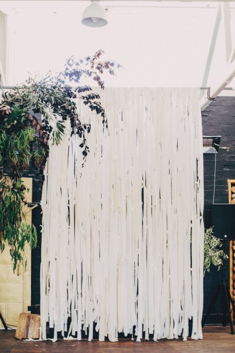 May include: A white backdrop made of fabric strips hangs against a black wall. The fabric strips are long and flowing, creating a soft and ethereal look. Greenery is draped over the top of the backdrop, adding a touch of natural beauty.