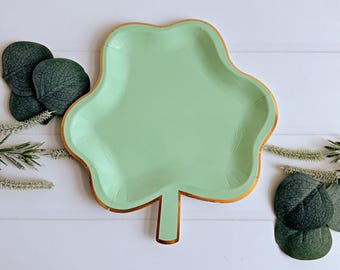 May include: A light green, leaf-shaped paper plate with a gold rim. The plate is surrounded by decorative greenery on a white wooden surface. The plate is approximately 10 inches long.
