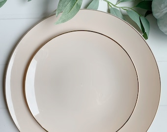 May include: A set of two stacked round plates in a soft cream color, each featuring a thin, metallic gold rim. The plates are smooth and glossy, with the smaller plate centered perfectly on top of the larger plate, creating a layered appearance.