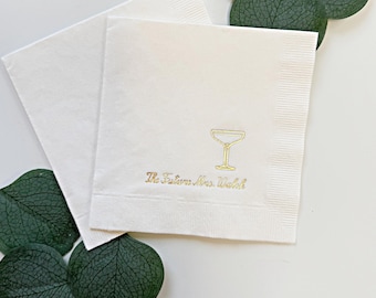 May include: Two square white paper napkins featuring a gold foil stamped martini glass icon and the text "The Future Mrs. Walsh" in a cursive script font. The napkins are layered and positioned with dark green eucalyptus leaves.