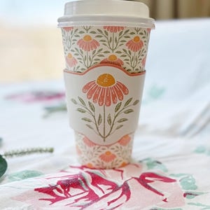 May include: A white disposable coffee cup with a floral design, featuring a matching sleeve and lid. The cup is adorned with a repeating pattern of pink flowers and green leaves. The sleeve showcases a large pink flower with a yellow center.