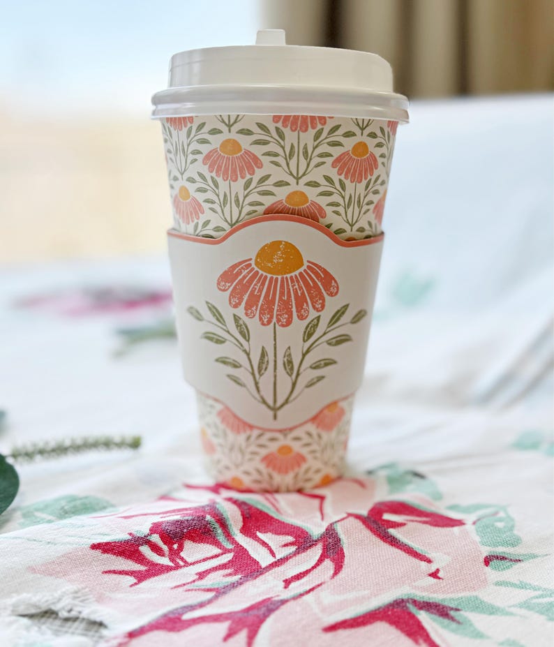 May include: A white disposable coffee cup with a floral design, featuring a matching sleeve and lid. The cup is adorned with a repeating pattern of pink flowers and green leaves. The sleeve showcases a large pink flower with a yellow center.