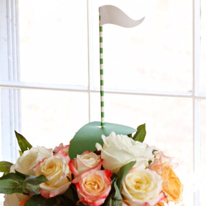 May include: A floral arrangement featuring a golf-themed centerpiece. The arrangement includes a variety of roses in shades of white, yellow, and peach. A green paper cutout simulates a golf green, with a striped green and white pole topped with a white flag.
