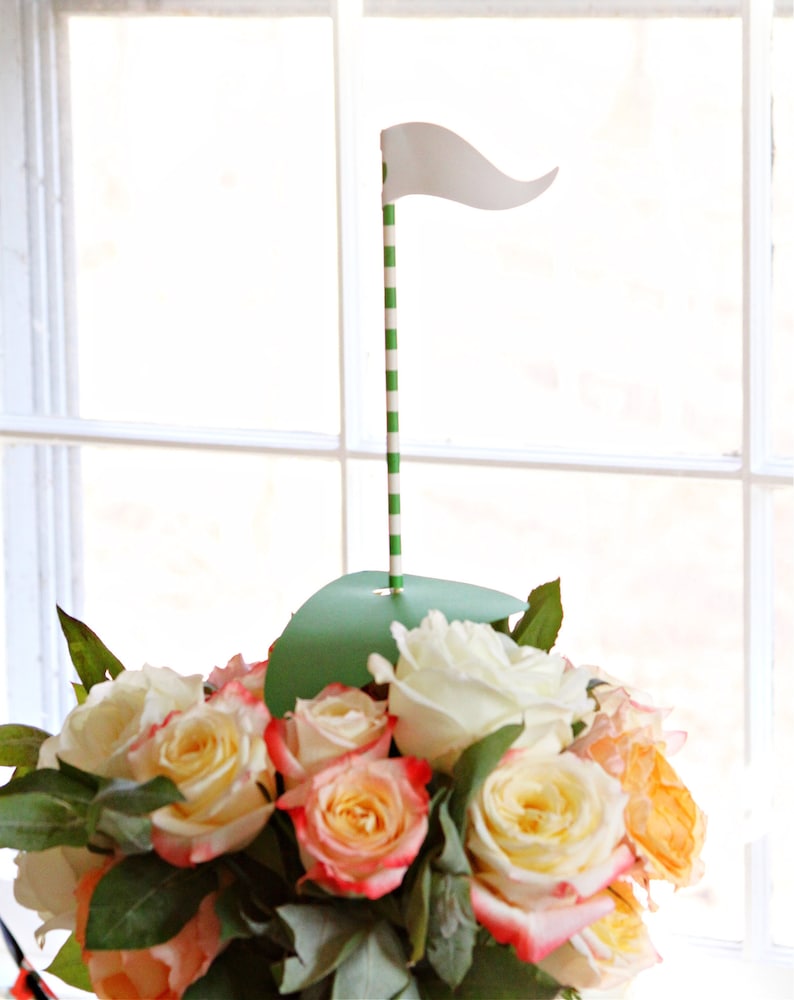 May include: A floral arrangement featuring a golf-themed centerpiece. The arrangement includes a variety of roses in shades of white, yellow, and peach. A green paper cutout simulates a golf green, with a striped green and white pole topped with a white flag.