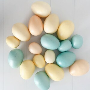 Op de afbeelding: Een bovenaanzicht toont een verzameling pastelkleurige paaseieren. De eieren vari&euml;ren in grootte en tint, waaronder lichtgeel, lichtroze en zachtblauw. Ze zijn gerangschikt op een wit oppervlak met subtiele verticale lijnen, wat een eenvoudige, strakke esthetiek cre&euml;ert.