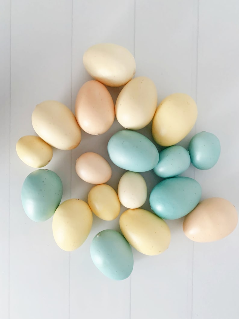 Op de afbeelding: Een bovenaanzicht toont een verzameling pastelkleurige paaseieren. De eieren vari&euml;ren in grootte en tint, waaronder lichtgeel, lichtroze en zachtblauw. Ze zijn gerangschikt op een wit oppervlak met subtiele verticale lijnen, wat een eenvoudige, strakke esthetiek cre&euml;ert.