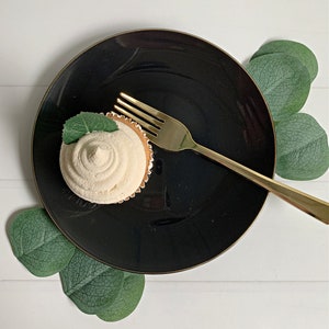 Op de afbeelding: Een cupcake met witte glazuur en een muntblad ligt op een zwart bord met een gouden rand. Een gouden vork ligt op het bord. Groene bladeren zijn rond het bord gerangschikt op een wit houten oppervlak.