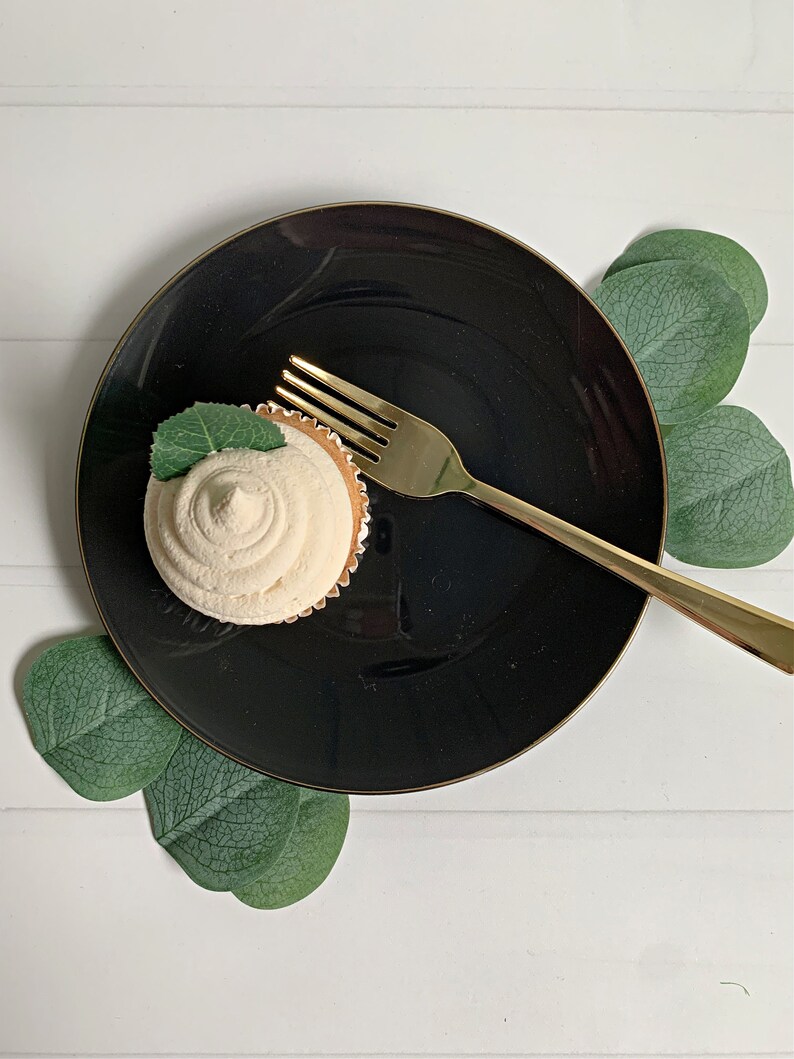 Op de afbeelding: Een cupcake met witte glazuur en een muntblad ligt op een zwart bord met een gouden rand. Een gouden vork ligt op het bord. Groene bladeren zijn rond het bord gerangschikt op een wit houten oppervlak.