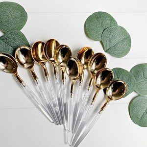 May include: A set of ten gold plastic spoons with clear plastic handles. The spoons are arranged in a fan shape on a white surface.