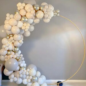 May include: A gold metal circular arch decorated with a cascade of white, cream, and silver balloons. The balloons are arranged in clusters and varying sizes, creating a festive and elegant display. Perfect for a wedding or party.