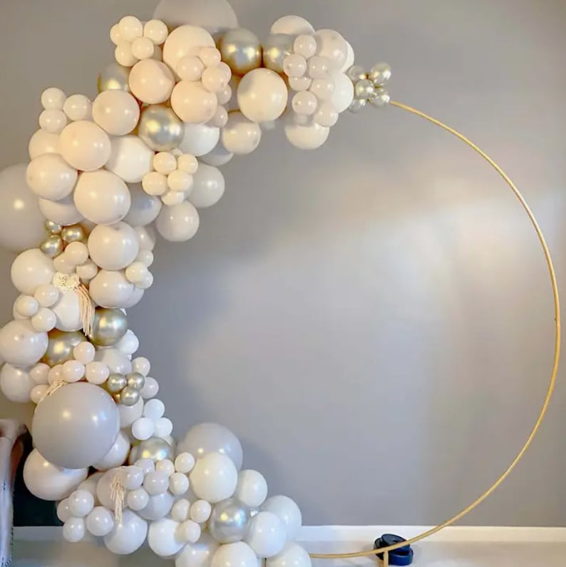 May include: A gold metal circular arch decorated with a cascade of white, cream, and silver balloons. The balloons are arranged in clusters and varying sizes, creating a festive and elegant display. Perfect for a wedding or party.