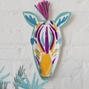 K&ouml;nnte beinhalten: Dekorative Zebra-Kopf-Dekoration aus Papier, mit wei&szlig;er Basis und lila, gelben und blauen Streifen. Es hat eine t&uuml;rkisfarbene Umrandung und einen bunten Fransen am oberen Rand. Das Kunstwerk ist an einer wei&szlig;en Ziegelmauer befestigt.
