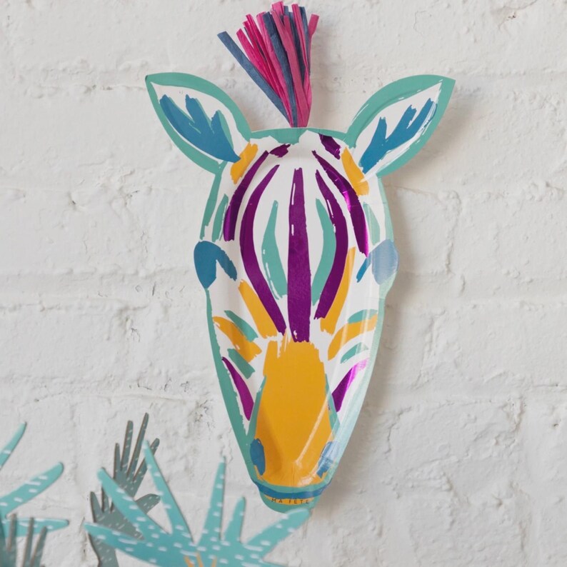 K&ouml;nnte beinhalten: Dekorative Zebra-Kopf-Dekoration aus Papier, mit wei&szlig;er Basis und lila, gelben und blauen Streifen. Es hat eine t&uuml;rkisfarbene Umrandung und einen bunten Fransen am oberen Rand. Das Kunstwerk ist an einer wei&szlig;en Ziegelmauer befestigt.