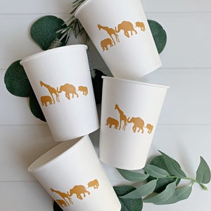 May include: Four white paper cups with gold animal silhouettes. The cups feature a giraffe, elephant, and rhino. The cups are arranged on a white surface with green leaves.
