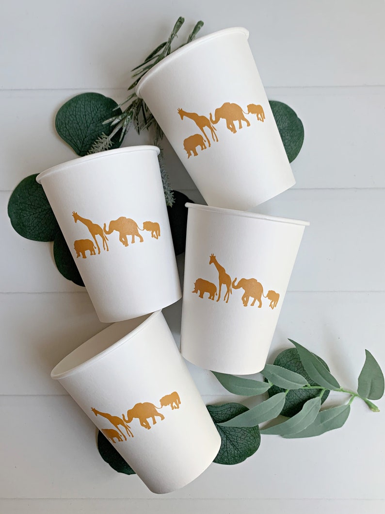 May include: Four white paper cups with gold animal silhouettes. The cups feature a giraffe, elephant, and rhino. The cups are arranged on a white surface with green leaves.
