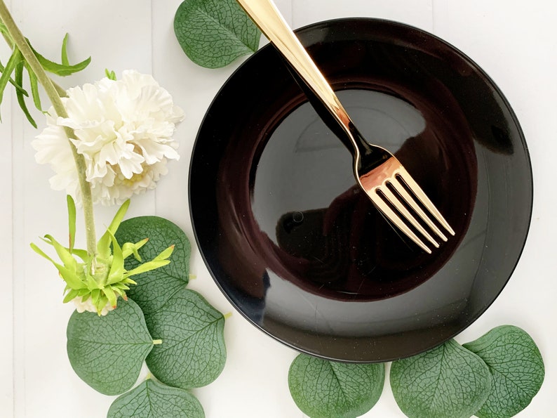 Puede incluir: Un plato negro con un tenedor dorado, rodeado de hojas verdes y una flor blanca. El tenedor est&aacute; colocado en diagonal sobre el plato. La escena est&aacute; ambientada sobre una superficie blanca, creando una est&eacute;tica limpia y elegante.