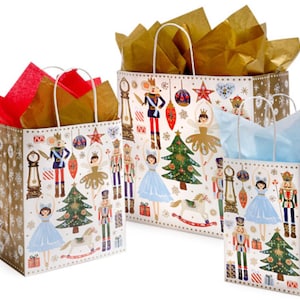 May include: Three gift bags with a Nutcracker theme. The bags are white with a gold snowflake pattern and feature illustrations of a Nutcracker, a ballerina, a Christmas tree, and other holiday elements. The bags are different sizes and come with tissue paper.