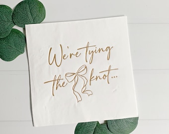 May include: White paper napkin with gold lettering that reads "We're tying the knot..." with a gold bow illustration.