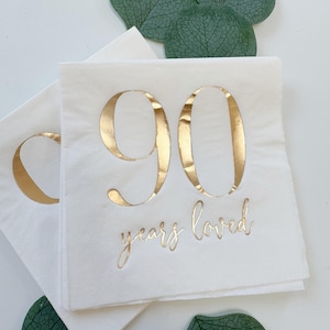 May include: White paper napkins featuring the number 90 in large, metallic gold foil lettering. Below the number, the text "years loved" is printed in a smaller, elegant metallic gold script font. The napkins are square with a smooth finish.