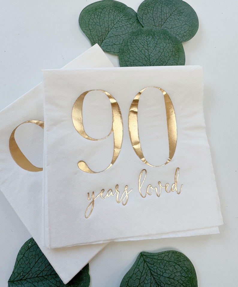 May include: White paper napkins featuring the number 90 in large, metallic gold foil lettering. Below the number, the text "years loved" is printed in a smaller, elegant metallic gold script font. The napkins are square with a smooth finish.
