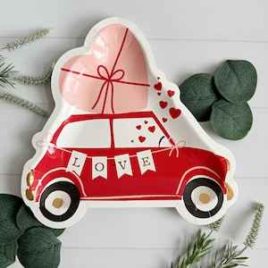 May include: Red paper party plate with a cartoon car carrying a pink heart-shaped gift box. The car has a banner that says "LOVE" and is surrounded by small red hearts.
