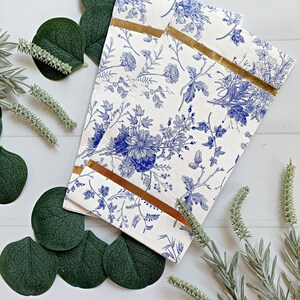 May include: Two white paper napkins with a blue floral pattern and gold trim. The napkins are folded in half and are lying on a white surface with green leaves and sprigs of lavender.