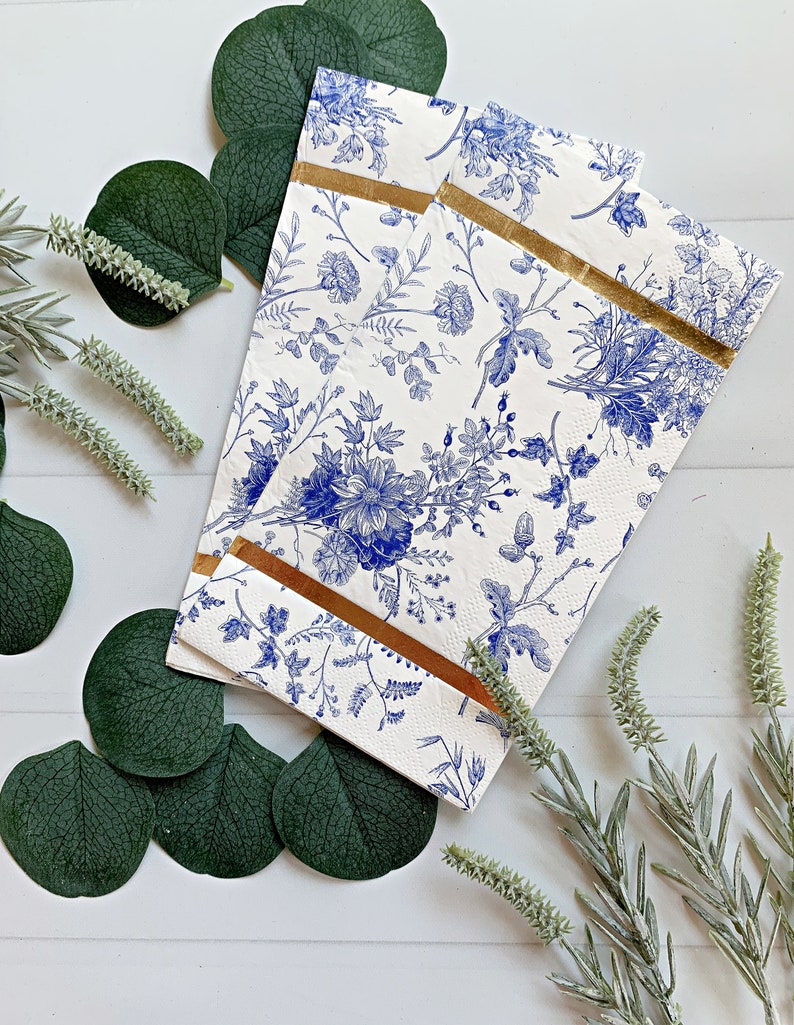May include: Two white paper napkins with a blue floral pattern and gold trim. The napkins are folded in half and are lying on a white surface with green leaves and sprigs of lavender.