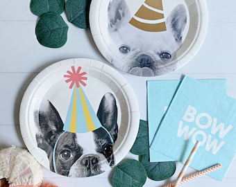 Può includere: Due piatti di carta bianchi con un'immagine in bianco e nero di un bulldog francese che indossa un cappello da festa. I piatti sono su una superficie di legno bianco con foglie verdi e cannucce a strisce arancioni e bianche. Un piatto ha un cappello da festa dorato e l'altro ha un cappello da festa blu e giallo. C'è un tovagliolo blu con il testo "BOW WOW" su di esso.