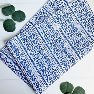 May include: Two folded white and blue patterned cloth napkins. The napkins feature a repeating design of blue shapes and lines. The napkins are placed on a white wooden surface, with green leaves in the corners.