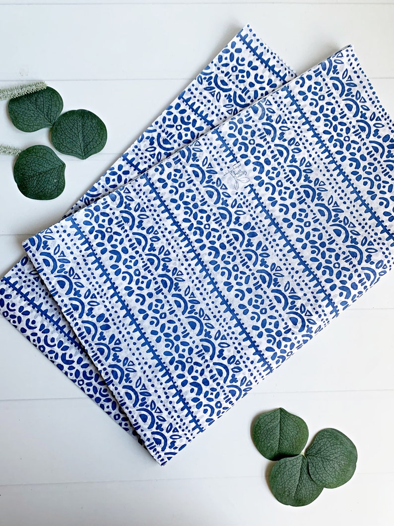 May include: Two folded white and blue patterned cloth napkins. The napkins feature a repeating design of blue shapes and lines. The napkins are placed on a white wooden surface, with green leaves in the corners.