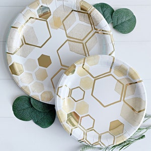 May include: Two white paper plates with a gold honeycomb pattern. The larger plate is behind the smaller plate. Both plates have a gold foil accent.