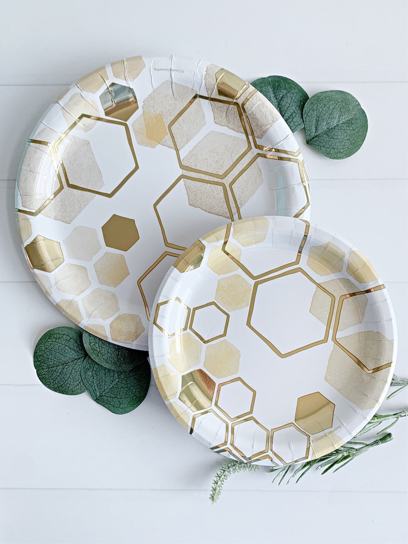 May include: Two white paper plates with a gold honeycomb pattern. The larger plate is behind the smaller plate. Both plates have a gold foil accent.