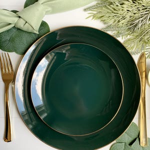 May include: Elegant dark green dinner plates with gold rims are arranged with gold silverware and a light green napkin. The table setting includes decorative greenery and eucalyptus leaves, creating a sophisticated and stylish presentation.
