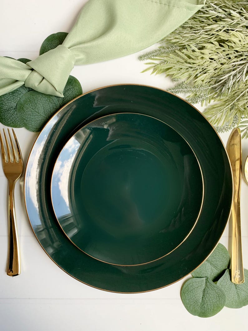 May include: Elegant dark green dinner plates with gold rims are arranged with gold silverware and a light green napkin. The table setting includes decorative greenery and eucalyptus leaves, creating a sophisticated and stylish presentation.