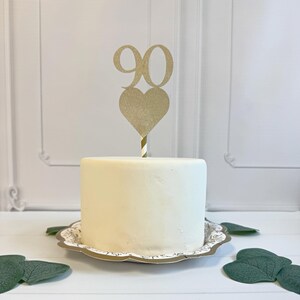 May include: A gold glitter cake topper featuring the number 90 positioned above a heart shape, attached to a striped stick. The topper is inserted into a small, smooth, white cylindrical cake resting on a decorative scalloped-edge plate.