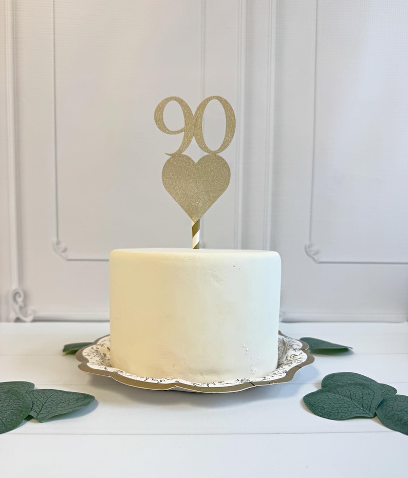 May include: A gold glitter cake topper featuring the number 90 positioned above a heart shape, attached to a striped stick. The topper is inserted into a small, smooth, white cylindrical cake resting on a decorative scalloped-edge plate.
