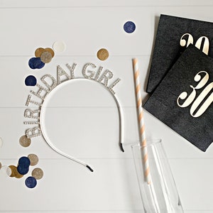 Puede incluir: Una diadema plateada con la inscripci&oacute;n "BIRTHDAY GIRL" con letras de pedrer&iacute;a, una copa de champ&aacute;n y una pajita a rayas. Servilletas negras con letras doradas "30" y confeti dorado, azul marino y blanco sobre una superficie de madera blanca.