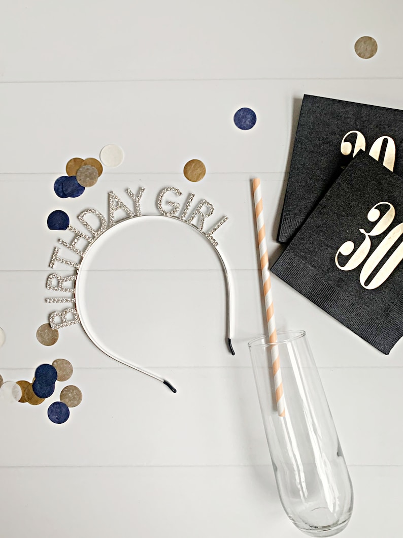 Puede incluir: Una diadema plateada con la inscripci&oacute;n "BIRTHDAY GIRL" con letras de pedrer&iacute;a, una copa de champ&aacute;n y una pajita a rayas. Servilletas negras con letras doradas "30" y confeti dorado, azul marino y blanco sobre una superficie de madera blanca.